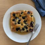 A slice of Sheet Pan Pancake with blueberries and maple syrup on a white plate