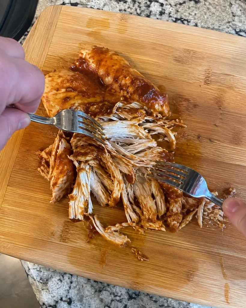 Shredding Crockpot BBQ Chicken with two forks on a cutting board