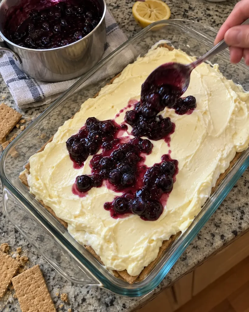 Assembling blueberry lemon icebox cake by spooning blueberry compote over lemon filling and graham crackers