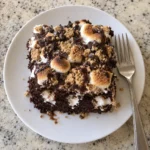 Single slice of s'mores poke cake on a white plate showing the marshmallow filling and toasted topping