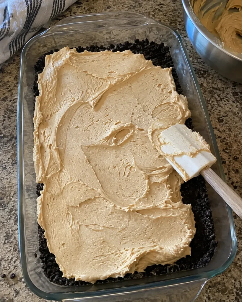 Spreading creamy peanut butter filling over the Oreo crust in a 9×13 pan Title: Spreading the Peanut Butter Layer